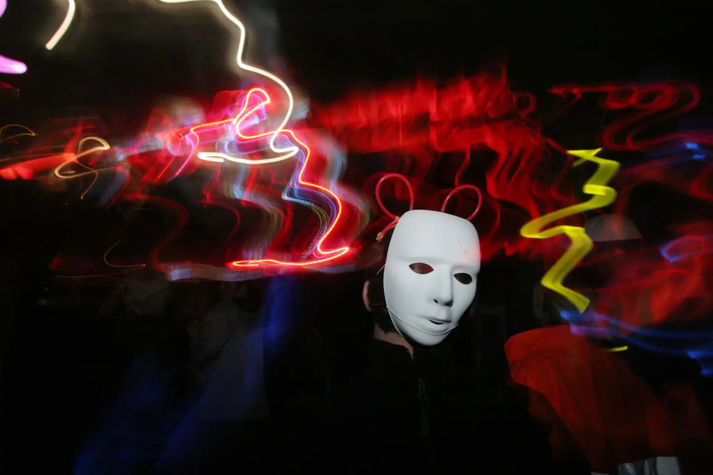 A dancer in a white mask under red and yellow light, the crowd blurred behind.