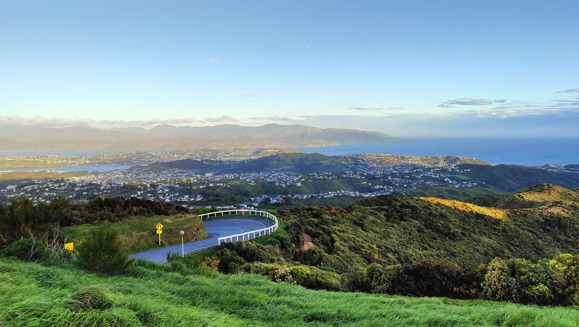 From the Brooklyn wind turbine: a road winding down through green hills toward the houses of Brooklyn and the south coast beyond, under a clear sky.